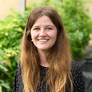 Charlotte Wright: Face and shoulders of lady with a big smile, in a garden. Long brown hair.
